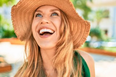 Yaz şapkası takmış, mutlu bir şekilde gülümseyen güzel sarışın bir kadın. Şehrin caddesinde yüzünde gülümsemeyle dikilirken.