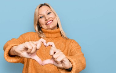 Orta yaşlı, beyaz kadın, sıradan bir kazak giyiyor, aşıkken gülümsüyor, elleriyle kalp sembolü yapıyor. romantik kavram. 