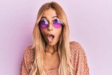 Güzel, sarışın, pembe güneş gözlüğü takan, ağzı açık, korkmuş, şaşırmış genç bir kadın. 