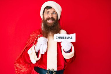 Uzun sakallı, yakışıklı, Noel Baba kostümü giyen, yüzünde mutlu ve havalı bir gülümsemeyle gülümseyen genç bir adam. diş gösterme. 