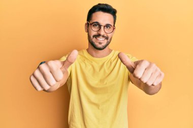 Gündelik kıyafetler giyen ve gözlük takan genç İspanyol adam elinle olumlu jest yapıyor, gülümsüyor ve başarı için mutlu. kazanan jest. 