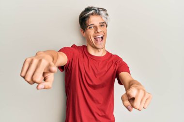 Gündelik kıyafetler giyen genç İspanyol adam seni ve kamerayı parmaklarıyla göstererek mutlu ve mutlu gülümsüyor. 