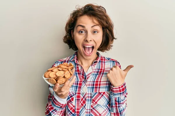 Young Brunette Woman Holding Bowl Salty Biscuits Pointing Thumb Side — Stock Photo, Image