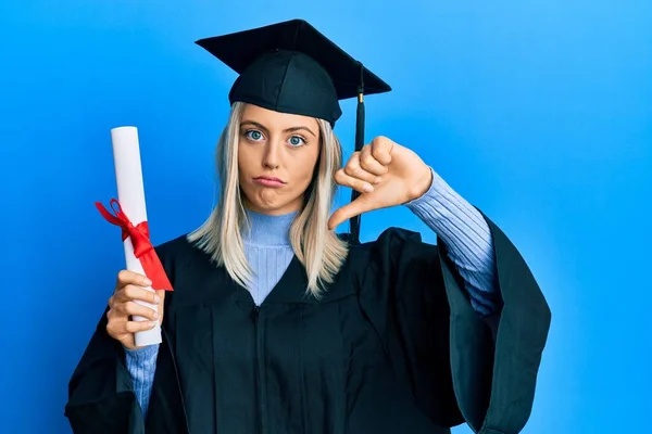 卒業キャップと式のローブを身に着けている美しいブロンドの女性怒り顔で学位を保持します 親指ダウンで嫌いを示す負の記号 拒絶概念 — ストック写真