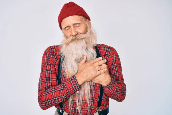 Old senior man with grey hair and long beard wearing hipster look with wool cap smiling with hands on chest with closed eyes and grateful gesture on face. health concept.