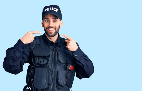 警官の制服を着た髭を生やした若いハンサムな男が陽気な笑みを浮かべて指の歯と口を指差している 歯の健康の概念 — ストック写真