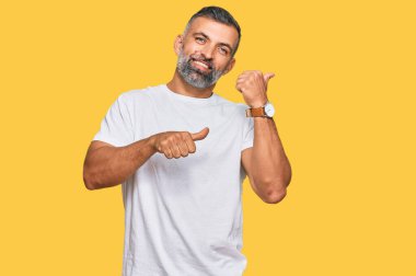 Orta yaşlı beyaz tişört giyen yakışıklı bir adam arka tarafı işaret ediyor. Elleri havada, kendinden emin bir şekilde gülümsüyor. 