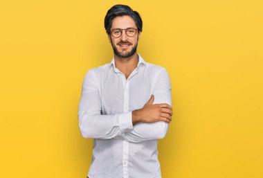 İş gömleği ve gözlük takan genç İspanyol adam kameraya bakarken kollarını kavuşturup gülümsüyor. pozitif kişi. 