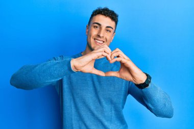Gündelik bir kazak giyen İspanyol genç adam, aşıkken gülümsüyor, elleriyle kalp sembolü yapıyor. romantik kavram. 