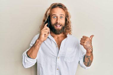 Sakallı ve uzun saçlı yakışıklı bir adam akıllı telefondan konuşuyor başparmağını kaldırıyor ve ağzı açık gülümsüyor. 