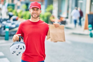 Beyaz bir teslimatçı, elinde kağıt torbayla şehirde mutlu bir şekilde gülümsüyor..
