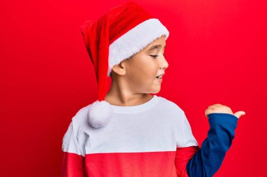 Küçük İspanyol çocuk Noel şapkası takıyor, gülümsüyor, gülümsüyor ve başparmağıyla yan tarafa bakıyor.. 