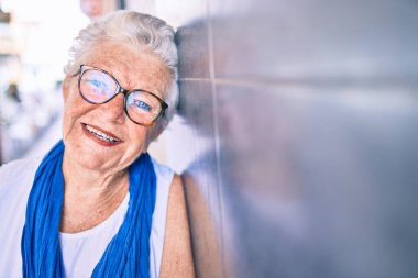 Yaşlı, yaşlı, kır saçlı, gülümseyen, açık havada duvara yaslanan yaşlı bir kadın.