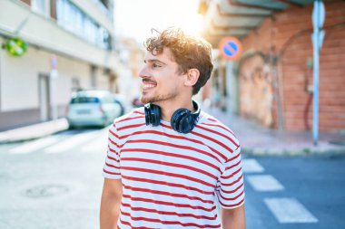 Genç beyaz adam mutlu bir şekilde gülümsüyor. Kulaklık takıyor ve şehirde yürüyor..