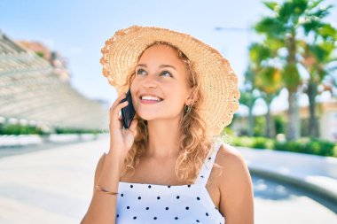 Sarışın, güzel, beyaz bir kadın. Dışarıda mutlu bir şekilde gülümsüyor. Telefonda konuşuyor.