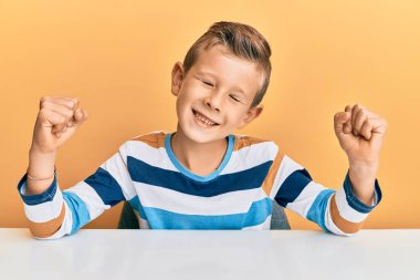 Sevimli beyaz çocuk, gündelik giysiler içinde masada mutlu ve heyecanlı bir şekilde oturuyor. Kazanan jestler yapıyor. Kolları havada, gülümsüyor ve başarı için bağırıyor. kutlama konsepti. 