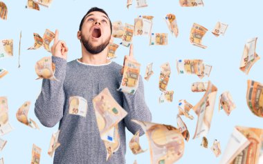 Gündelik süveter giyen genç yakışıklı adam şaşırmış ve şaşırmış bir şekilde yukarı bakıyor ve parmaklarıyla işaret ediyor ve kollarını kaldırıyor..