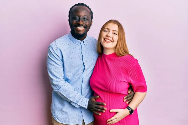 Young interracial couple expecting a baby, touching pregnant belly with a happy and cool smile on face. lucky person.