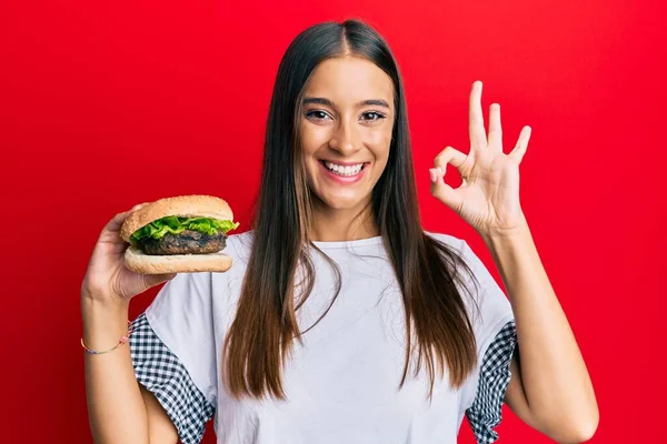 Fiatal Spanyol Eszik Hamburger Csinál Rendben Jel Ujjaival Mosolygós Barátságos — Stock Fotó