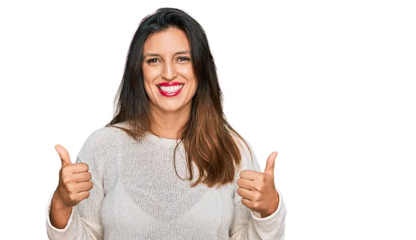 手で積極的なジェスチャーを行うカジュアルセーターの成功サインを身に着けている美しいヒスパニック系の女性 笑顔と幸せを親指アップ 陽気な表情と勝者のジェスチャー — ストック写真