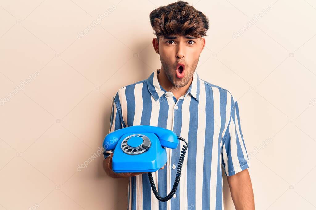Young hispanic man holding vintage telephone scared and amazed with open mouth for surprise, disbelief face 