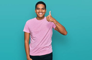 Young handsome hispanic man wearing casual pink t shirt smiling happy and positive, thumb up doing excellent and approval sign 