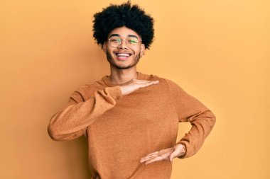 Young african american man with afro hair wearing casual winter sweater gesturing with hands showing big and large size sign, measure symbol. smiling looking at the camera. measuring concept. 
