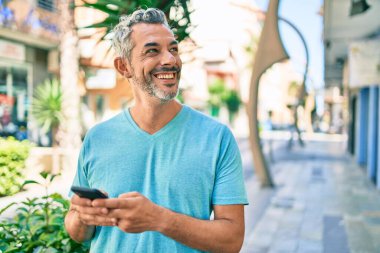 Orta yaşlı, kır saçlı, mutlu gülümseyen bir adam şehrin caddesinde yürürken akıllı telefon kullanıyor..