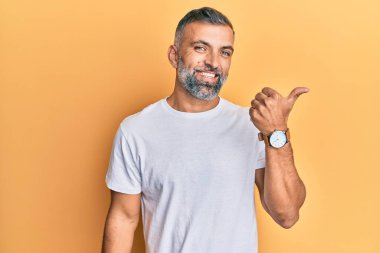 Orta yaşlı, beyaz tişört giyen, mutlu yüzlü, gülümseyen ve baş parmağıyla yan tarafı gösteren yakışıklı bir adam.. 
