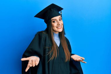 Mezuniyet şapkası ve tören cübbesi giyen güzel esmer bir kadın. Kolları ve elleri havada, kafası karışmış bir ifade takınıyor. şüphe kavramı. 