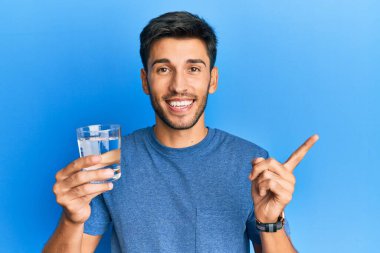 Genç yakışıklı adam, bir bardak su içerken gülümsüyor mutlu bir şekilde elini ve parmağını yana doğru gösteriyor. 