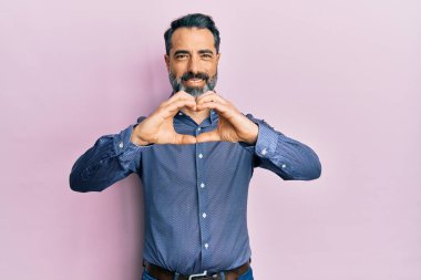 Orta yaşlı, sakallı, gri saçlı, iş kıyafetleri giyen, aşkta gülümseyen, elleriyle kalp sembolü yapan bir adam. romantik kavram. 