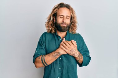 Sakallı, uzun saçlı, sıradan kıyafetler giyen, göğsünde elleri, yüzünde minnet dolu bir jestle gülümseyen yakışıklı bir adam. sağlık kavramı. 