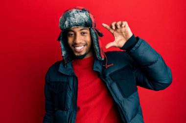 Sakallı, kışlık şapkalı, ceketli, gülümseyen, kendine güvenen, el hareketi yapan, parmaklarıyla işaret yapan ve kameraya bakan Afrikalı genç bir Amerikalı. ölçüm kavramı. 