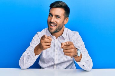 Masada oturmuş, mutlu ve komik yüzüyle kamerayı işaret eden iş kıyafetleri giymiş yakışıklı bir İspanyol. iyi enerji ve titreşimler. 