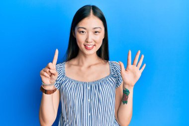 Genç Çinli kadın, sıradan çizgili tişört giyip altı numaralı parmaklarıyla işaret ederken kendinden emin ve mutlu gülümsüyor.. 