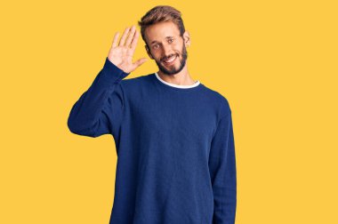 Sarışın, sakallı, sıradan bir süveter giyen, mutlu, güler yüzlü, dostça bir jest yapan yakışıklı bir adam. 