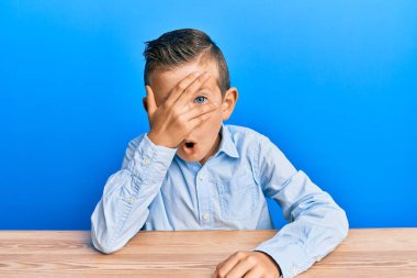 Sevimli beyaz çocuk, günlük giysiler içinde masaya oturmuş, şok içinde yüzünü ve gözlerini eliyle kapatmış, parmaklarından korkuyla bakıyor. 
