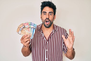Handsome hispanic man with beard holding canadian dollars scared and amazed with open mouth for surprise, disbelief face 