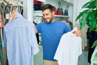 Genç, yakışıklı, gülümseyen, giyim mağazasında giysileri tutan.