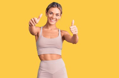 Beyaz giyimli güzel bir kadın spor kıyafetleri giyiyor. Elleriyle olumlu bir jest yapıyor. Gülümsüyor ve başarı için mutlu. kazanan jest. 