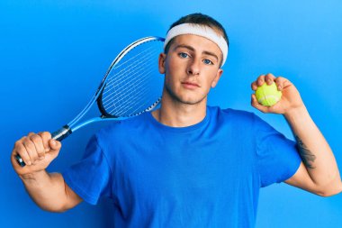 Tenis oynayan beyaz bir adam raket ve top tutuyor ve yüzünde ciddi bir ifadeyle rahatlıyor. Basit ve doğal bir şekilde kameraya bakıyorsun.. 