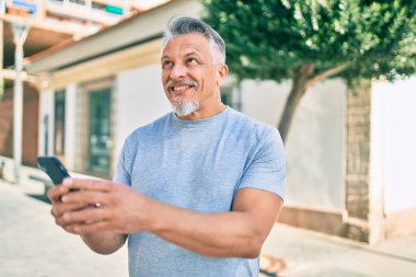 Orta yaşlı, ispanyol, kır saçlı, mutlu gülümseyen adam şehirde akıllı telefon kullanıyor..