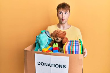 Young caucasian man holding donation box with toys making fish face with mouth and squinting eyes, crazy and comical. 