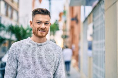 Genç beyaz adam gülümseyerek şehirde mutlu mesut duruyor..