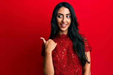 Güzel İspanyol kadın, kırmızı arka planda zarif kıyafetler giyip gülümsüyor, gülümsüyor ve başparmağıyla yan tarafı işaret ediyor.. 