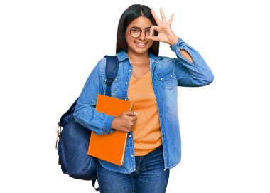 Sırt çantası takan, kitap tutan, el hareketleriyle gülümseyen, mutlu yüzüyle parmaklarının arasından bakan genç Latin kız.. 