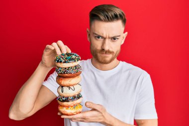 Genç kızıl saçlı adam elinde lezzetli renkli çörekler tutuyor şüpheci ve gergin, sorun yüzünden surat asıyor. negatif kişi. 