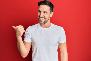 Genç, yakışıklı, günlük beyaz tişört giyen, baş parmağını yana doğru gösteren, ağzı açık gülümseyen bir adam. 
