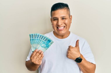 Genç Latin adam elinde 100 Brezilyalı gerçek banknot tutuyor mutlu ve pozitif gülümsüyor, başparmağıyla mükemmel ve onay işareti yapıyor. 